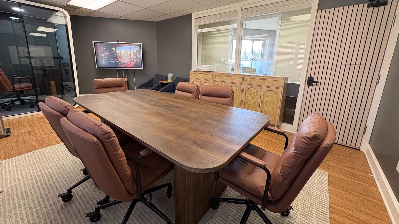 Harbor's conference room: a solid walnut table ringed with tan leather chairs, a wall-mounted screen and camera at the far end, and a glass phone booth through the doorway.