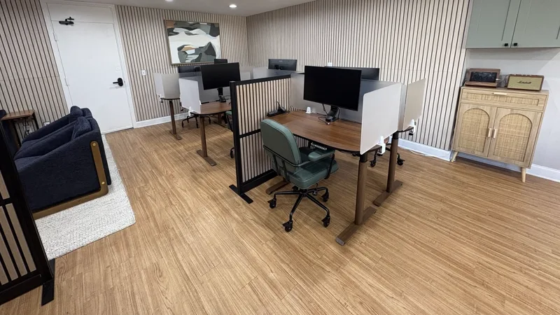 Sit-stand desks with privacy screens and green leather chairs against a slatted wood wall, with a sofa and rug at the far end.