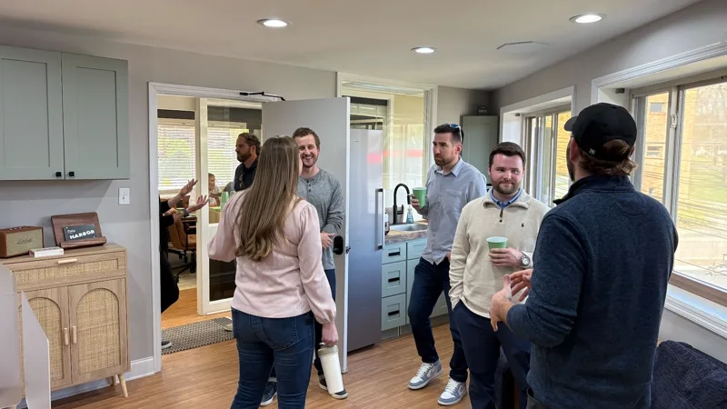 Members standing and talking with drinks in the kitchen area of Harbor, the Harbor sign on the credenza behind them.