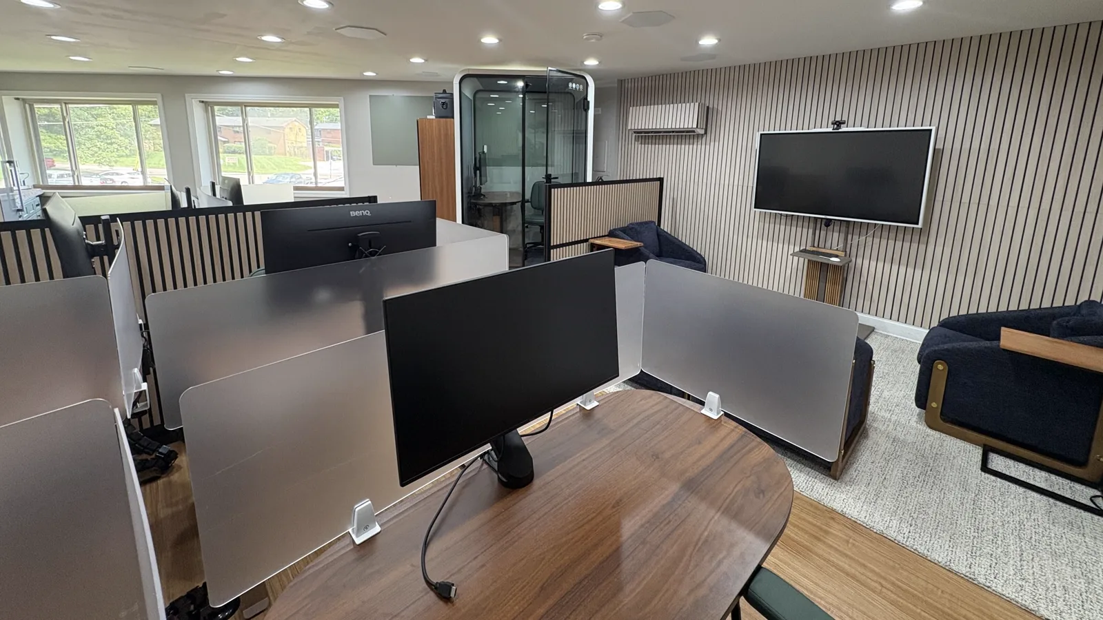 The main room at Harbor: sit-stand desks with privacy screens, an enclosed glass phone booth, a wall-mounted screen and daylight from the street-side windows.