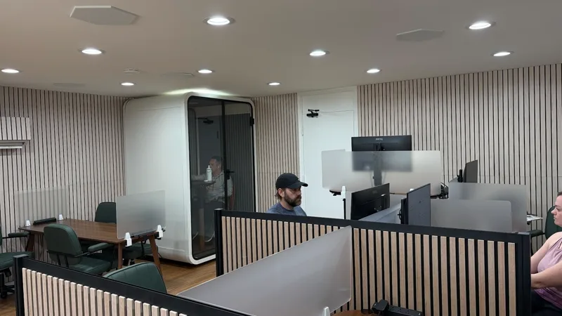 Three members at work in the main room: one inside the glass phone pod on a call, two at desks behind privacy screens.